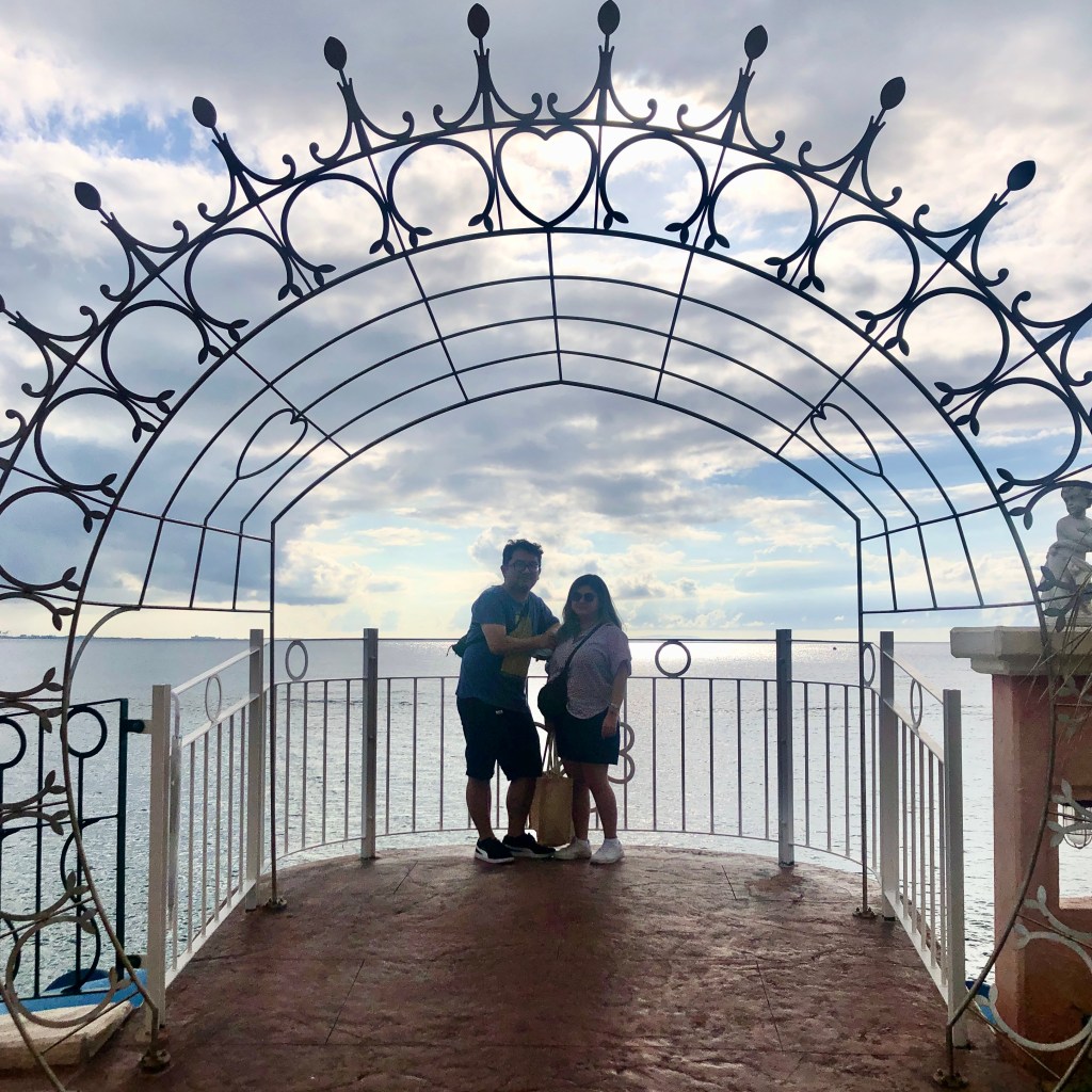 Casal posando numa espécie de varanda com grade bonita, na orla da praia, com chão de piso avermelhado e mar ao fundo. O céu está azul brilhante e o casal aparece pequeno e escuro na foto.